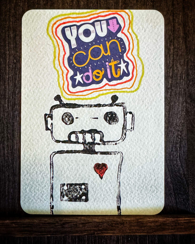 Selbstgemachte Postkarte mit einem Linocut-Print Roboter und einem Aufklaber mit dem Text "You can do it!".